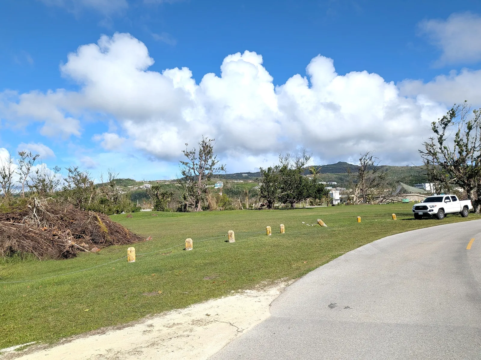 Piled storm debris on Micro Beach's green during ongoing typhoon cleanup, with Saipan's central hills in the background